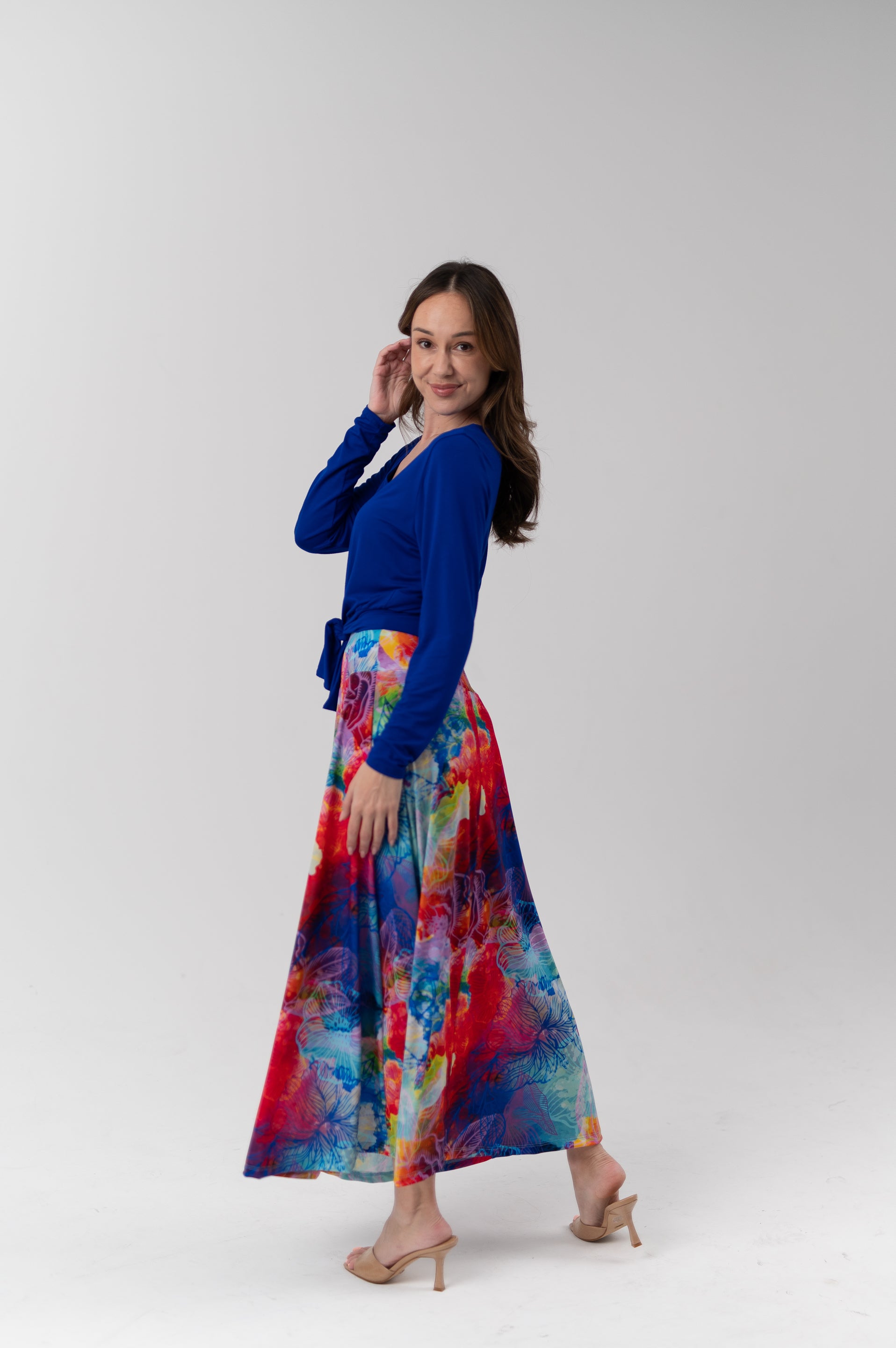 A woman modeling a vibrant, multicolored abstract-print maxi skirt paired with a royal blue long-sleeve top, standing against a plain light gray studio background.