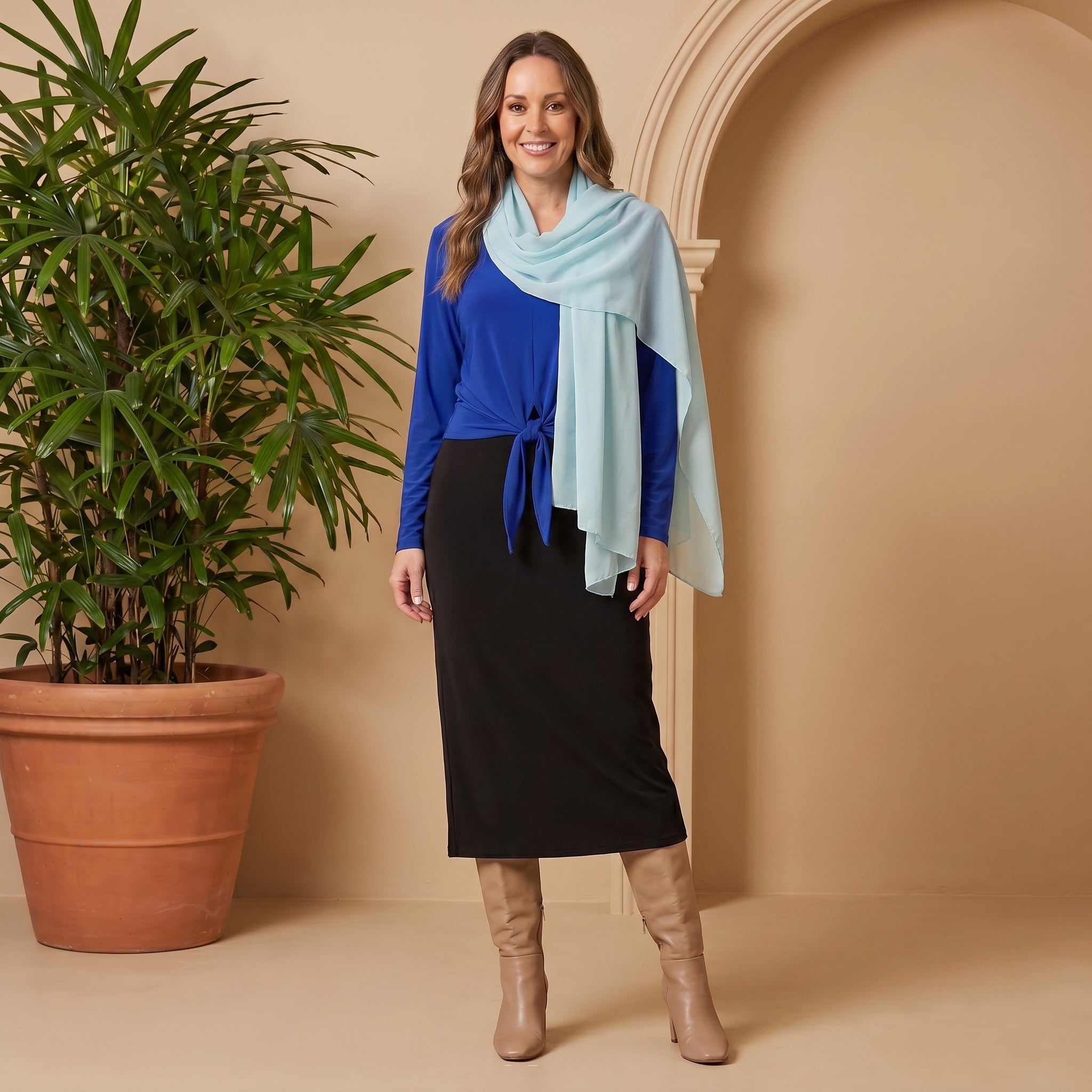 Woman wearing a blue top, black skirt, and beige boots standing next to a potted plant against a beige wall.