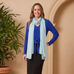 Woman in blue top and black skirt with light blue scarf standing next to a potted plant against a beige wall.