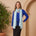 Woman in blue top and black skirt with light blue scarf standing next to a potted plant against a beige wall.