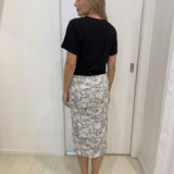 Woman wearing a black top and patterned skirt standing against a white wall.