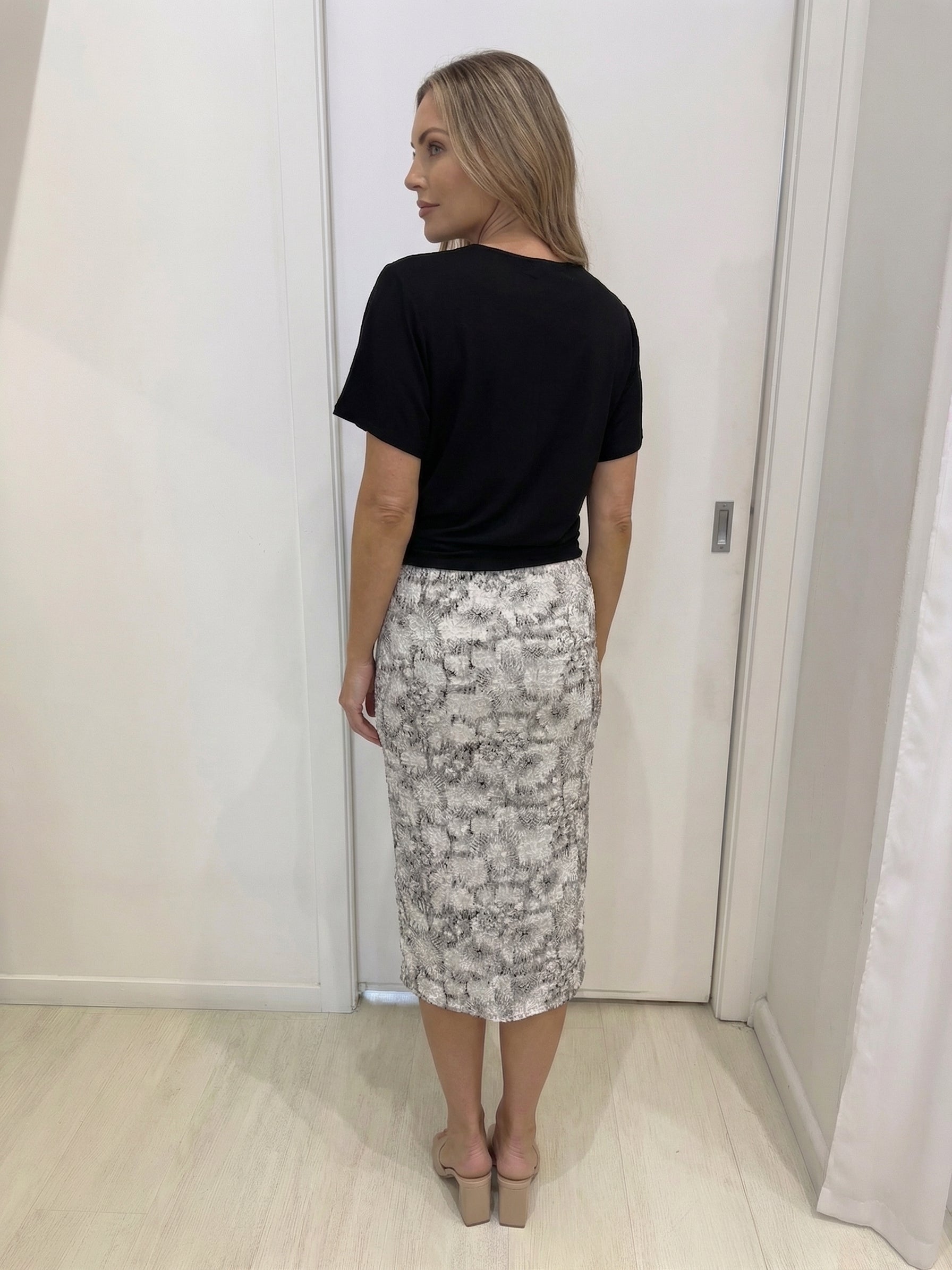 Woman wearing a black top and patterned skirt standing against a white wall.