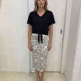 Woman wearing a black top and patterned skirt in a room with white walls and wooden floor.