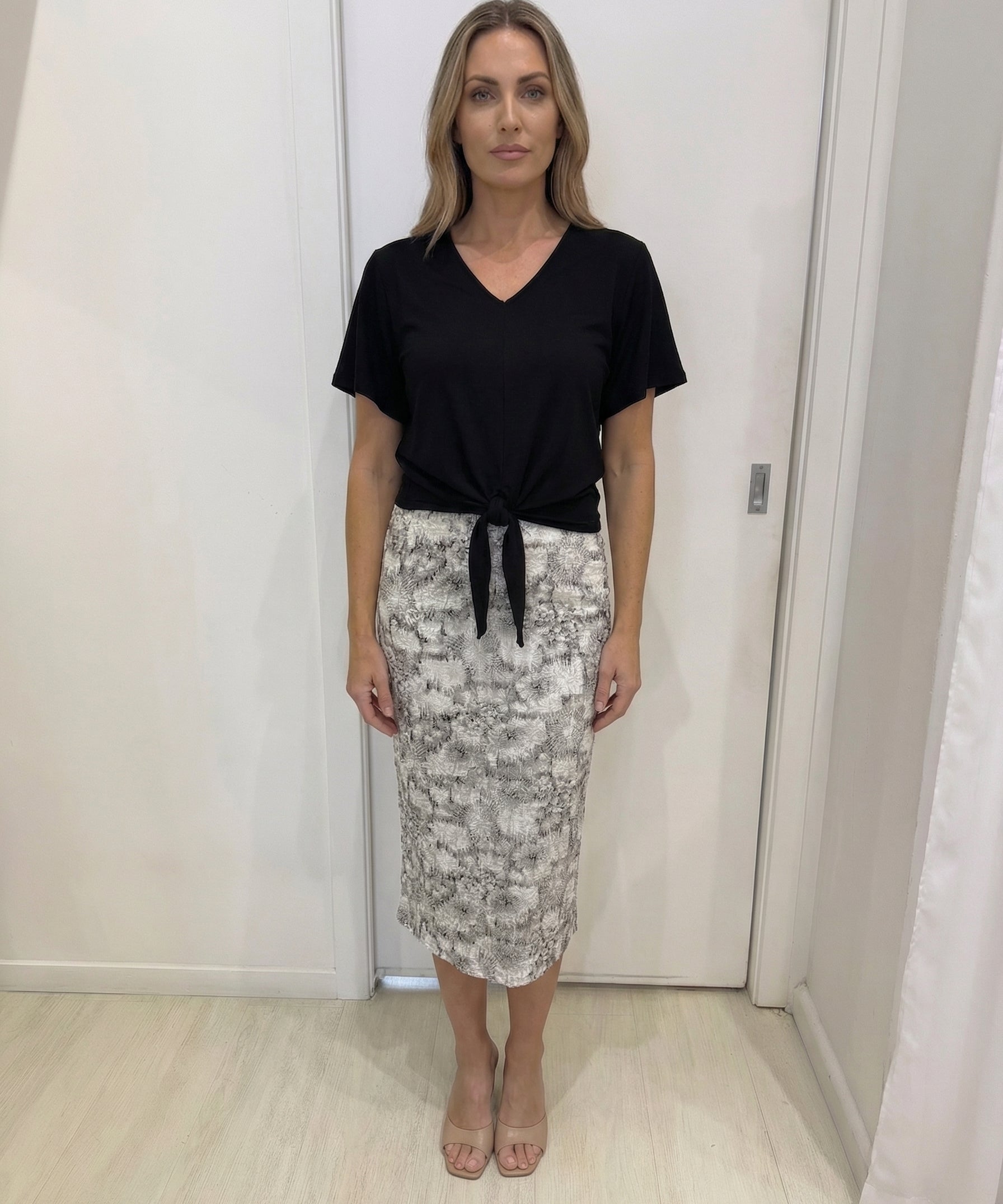 Woman wearing a black top and patterned skirt in a room with white walls and wooden floor.