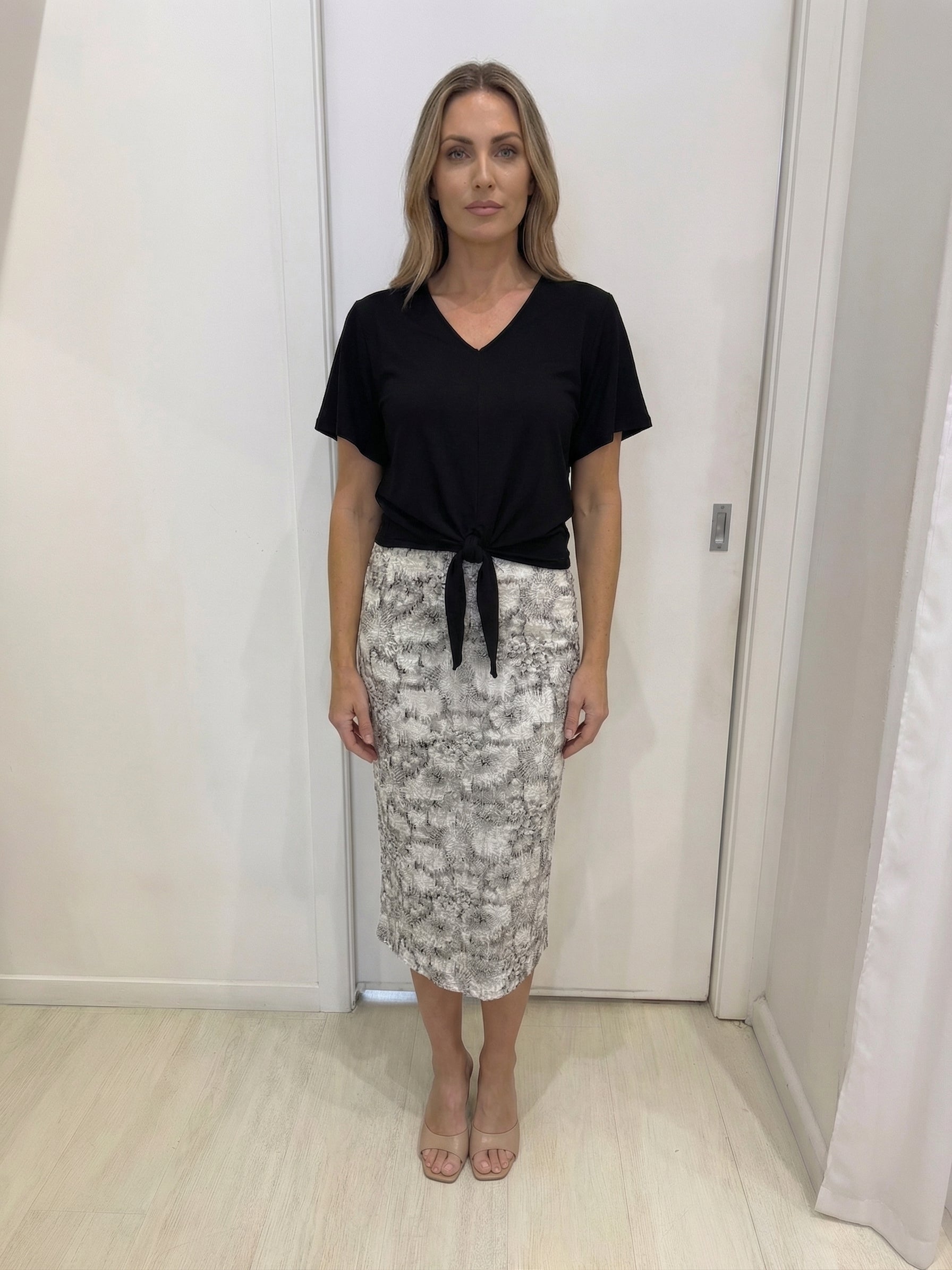 Woman wearing a black top and patterned skirt in a room with white walls and wooden floor.
