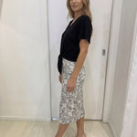 Woman wearing a black top and patterned skirt standing against a white wall.