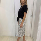 Woman wearing a black top and patterned skirt standing against a white wall.