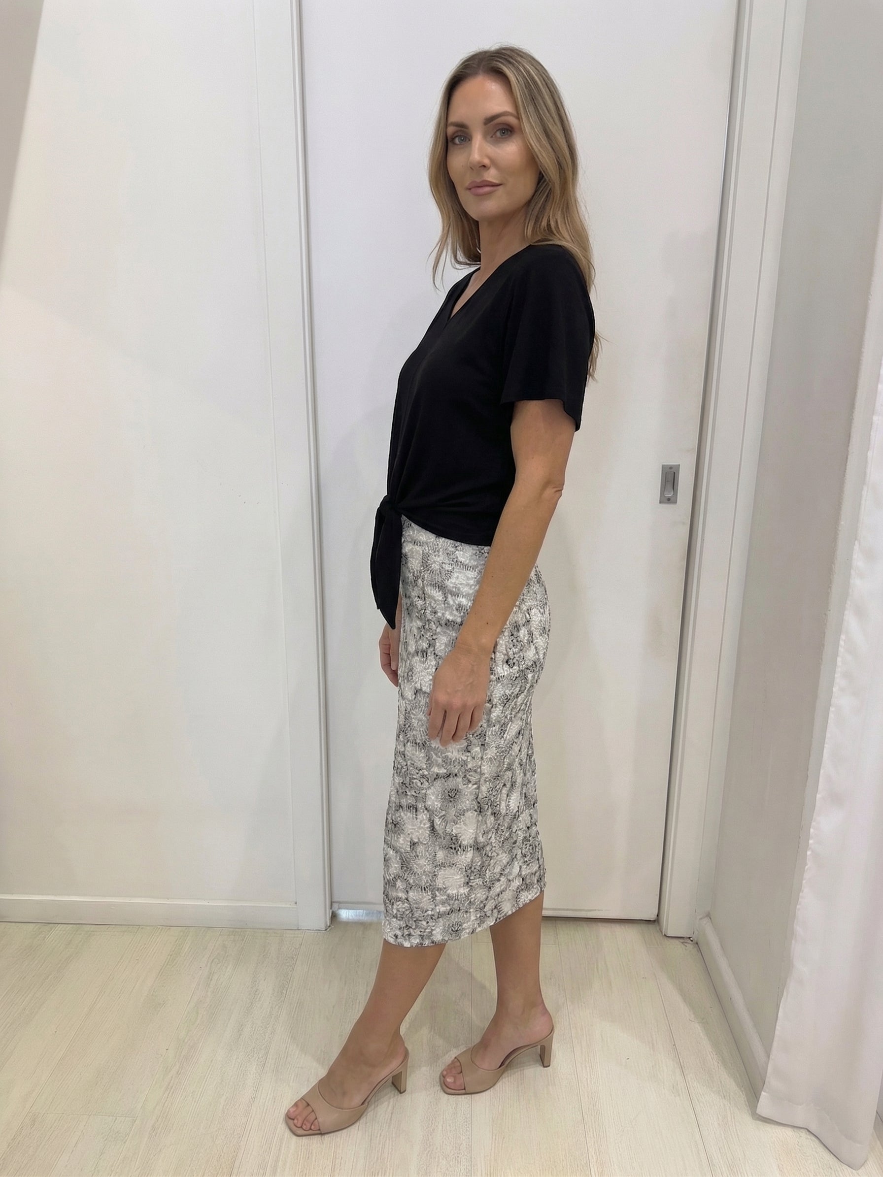 Woman wearing a black top and patterned skirt standing against a white wall.