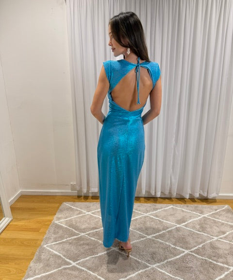 A woman wearing a shimmering turquoise blue maxi dress with a back tie detail, standing on a patterned rug in front of white curtains.