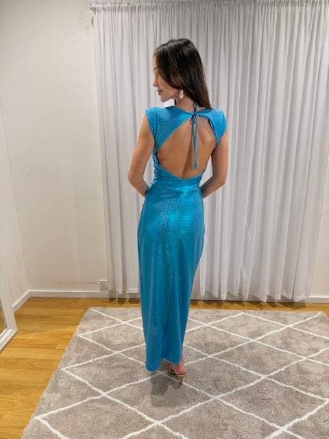 A woman wearing a shimmering turquoise blue maxi dress with a back tie detail, standing on a patterned rug in front of white curtains.