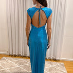 A woman stands with her back to the camera wearing a shimmering bright blue maxi dress with a tie-back open back, photographed indoors in front of white curtains and a patterned rug.