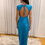 A woman stands with her back to the camera wearing a shimmering bright blue maxi dress with a tie-back open back, photographed indoors in front of white curtains and a patterned rug.