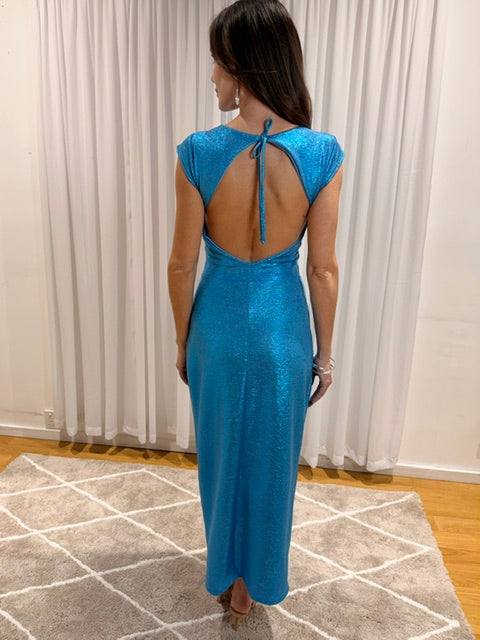 A woman stands with her back to the camera wearing a shimmering bright blue maxi dress with a tie-back open back, photographed indoors in front of white curtains and a patterned rug.