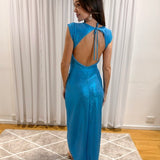 A woman wearing a sparkling blue sleeveless maxi dress with a large open back and tie detail, standing in a room with white curtains and a patterned rug.