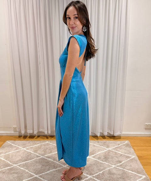 A woman modeling a sleeveless, backless turquoise blue maxi dress with a shimmering finish, standing on a patterned rug in front of white curtains.