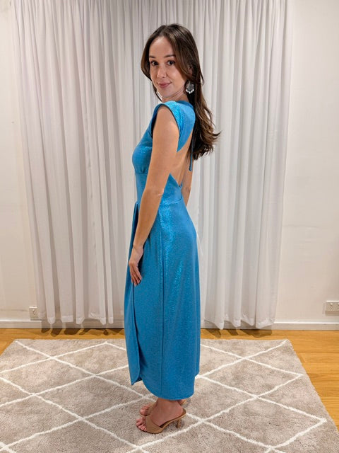 A woman modeling a sleeveless, backless turquoise blue maxi dress with a shimmering finish, standing on a patterned rug in front of white curtains.