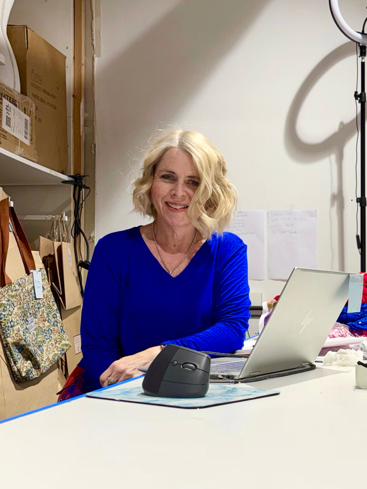 A smiling blonde woman in a vibrant royal blue long-sleeve top sits at a white desk with a laptop, computer mouse, and a ring light visible in the background of what looks like a workspace or studio.