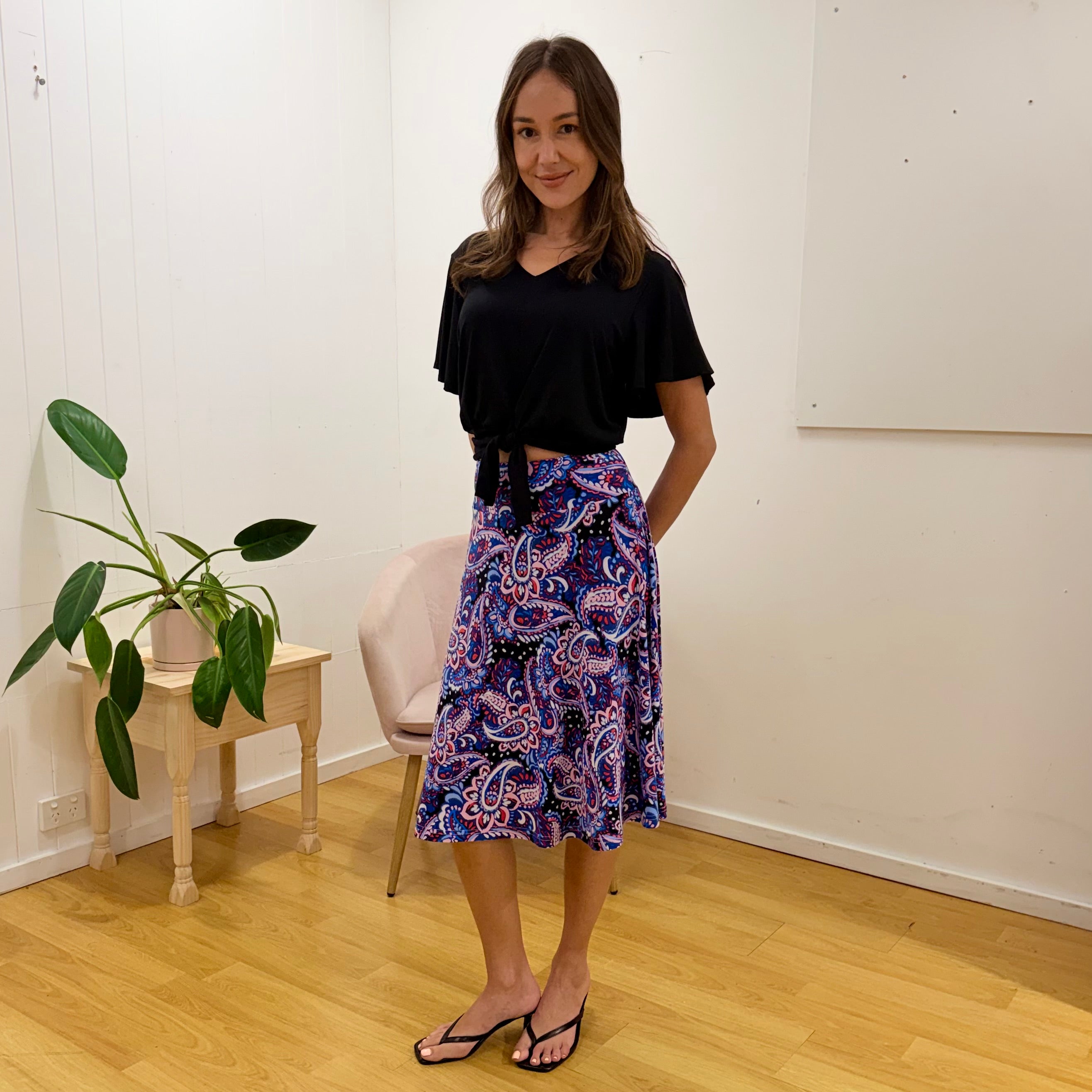 Woman wearing a black top and colorful patterned skirt standing in a room with a plant and chair.