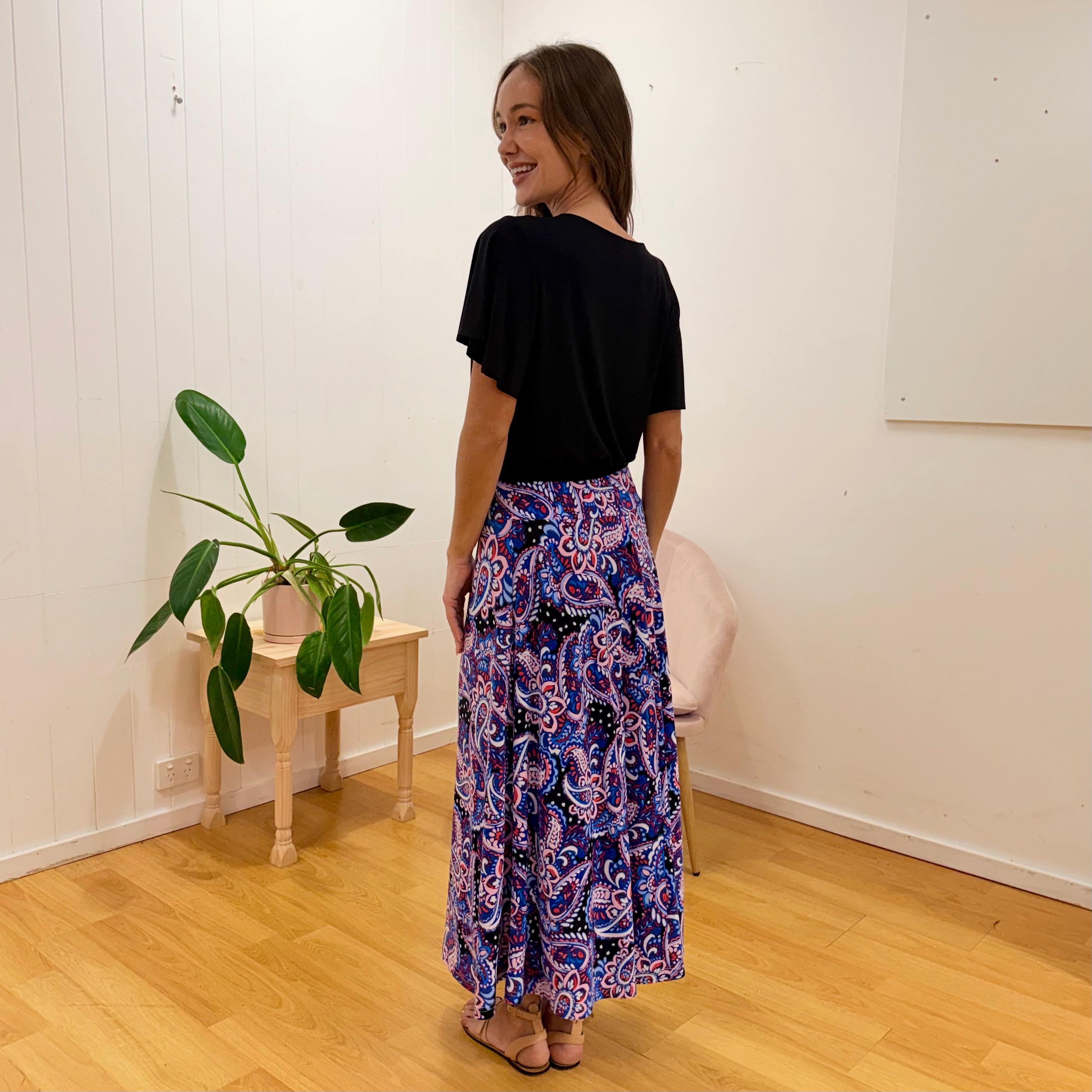 Woman wearing a black top and colorful patterned skirt standing in a room with a plant and wooden side table.