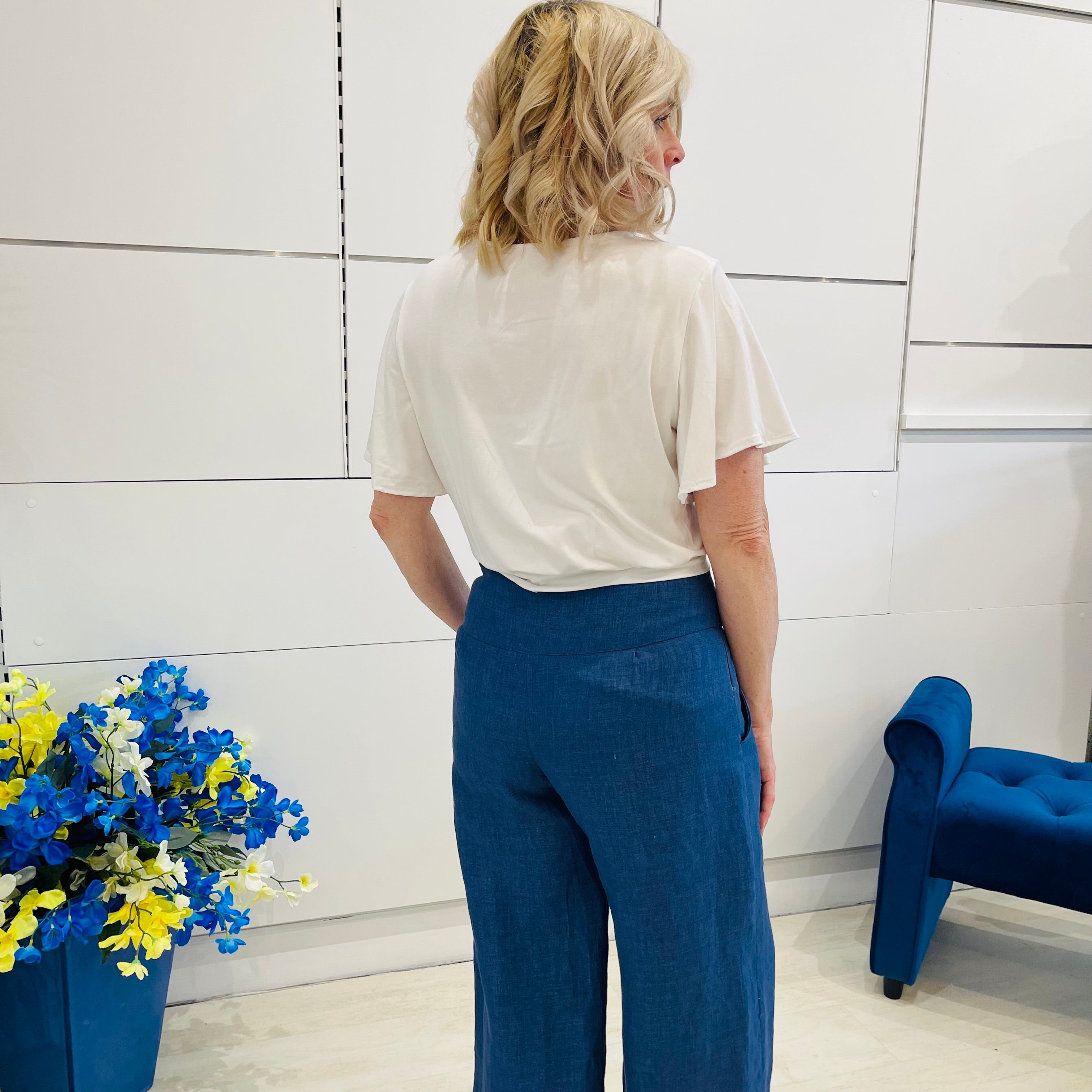Woman wearing a white top and blue pants standing in a room with a blue chair and floral arrangement.