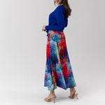 A woman modeling a vibrant, multicolored abstract print maxi skirt paired with a royal blue long-sleeve top, standing against a plain light gray studio background.
