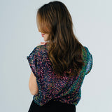 A woman shown from the back wearing a short-sleeve top covered in dense multicolor sequins, paired with a black skirt, against a plain light gray background.