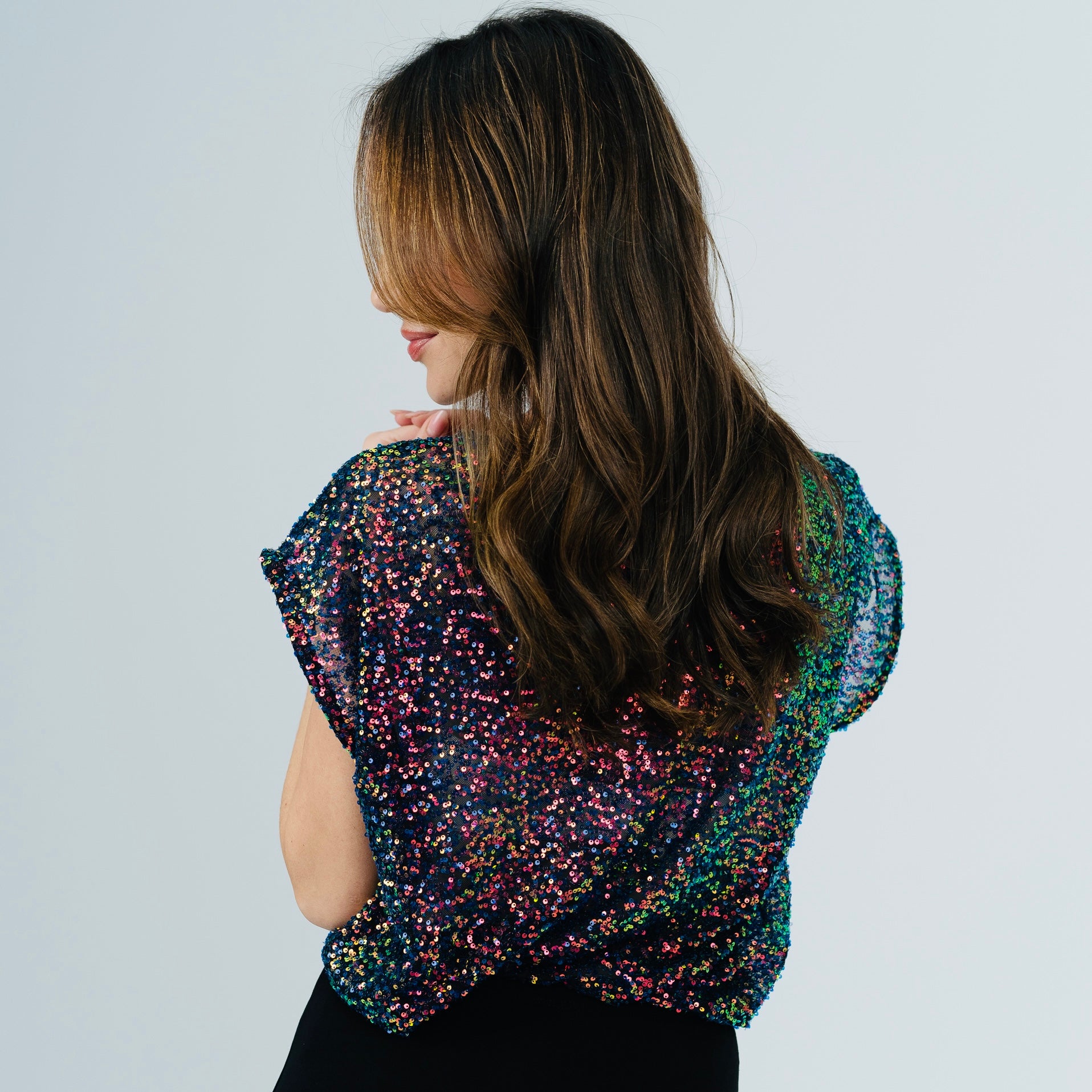 A woman shown from the back wearing a short-sleeve top covered in dense multicolor sequins, paired with a black skirt, against a plain light gray background.