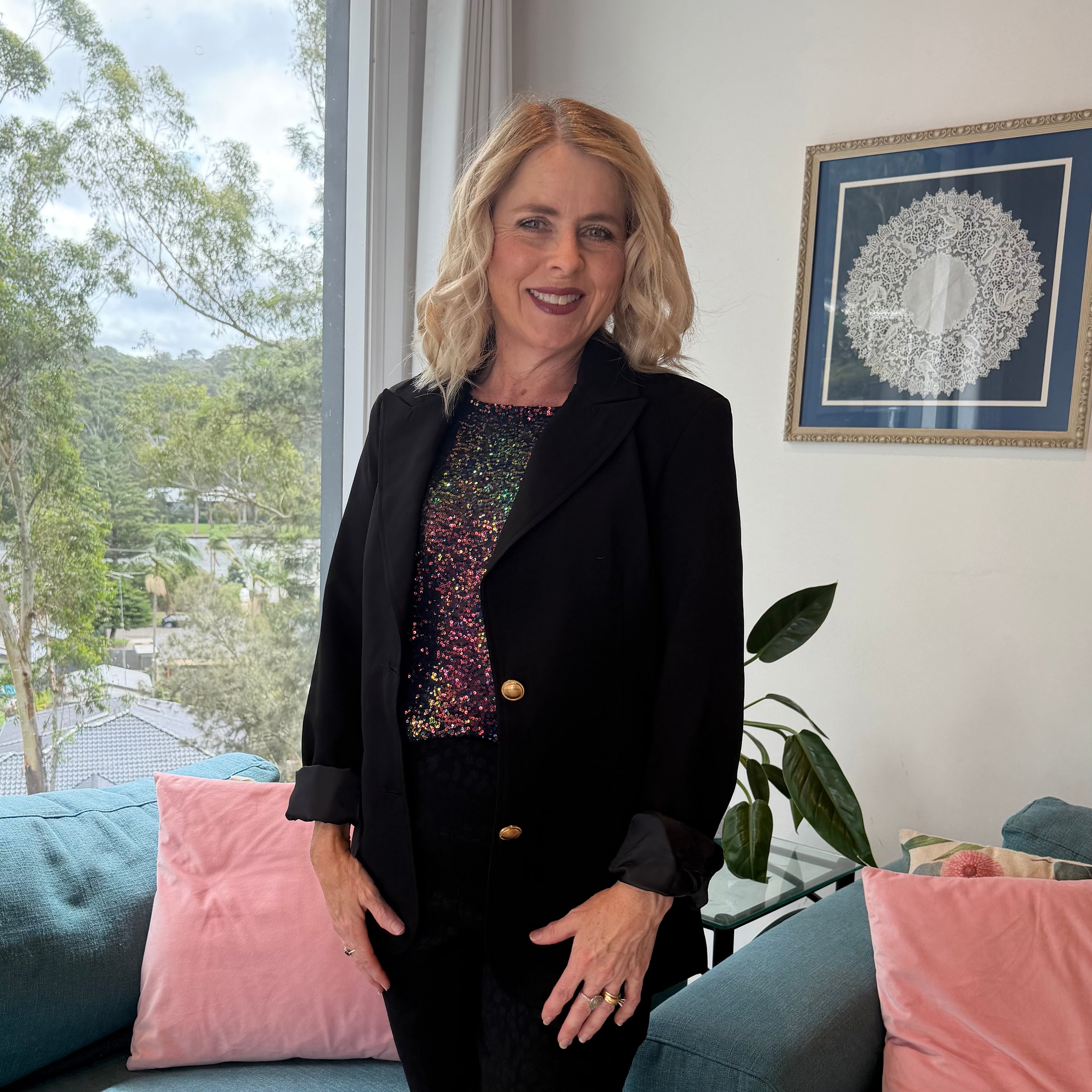 Woman in a black suit standing in a living room with pink cushions and a window view.