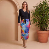 Woman wearing a black top and colorful skirt standing next to a potted plant against a beige wall.