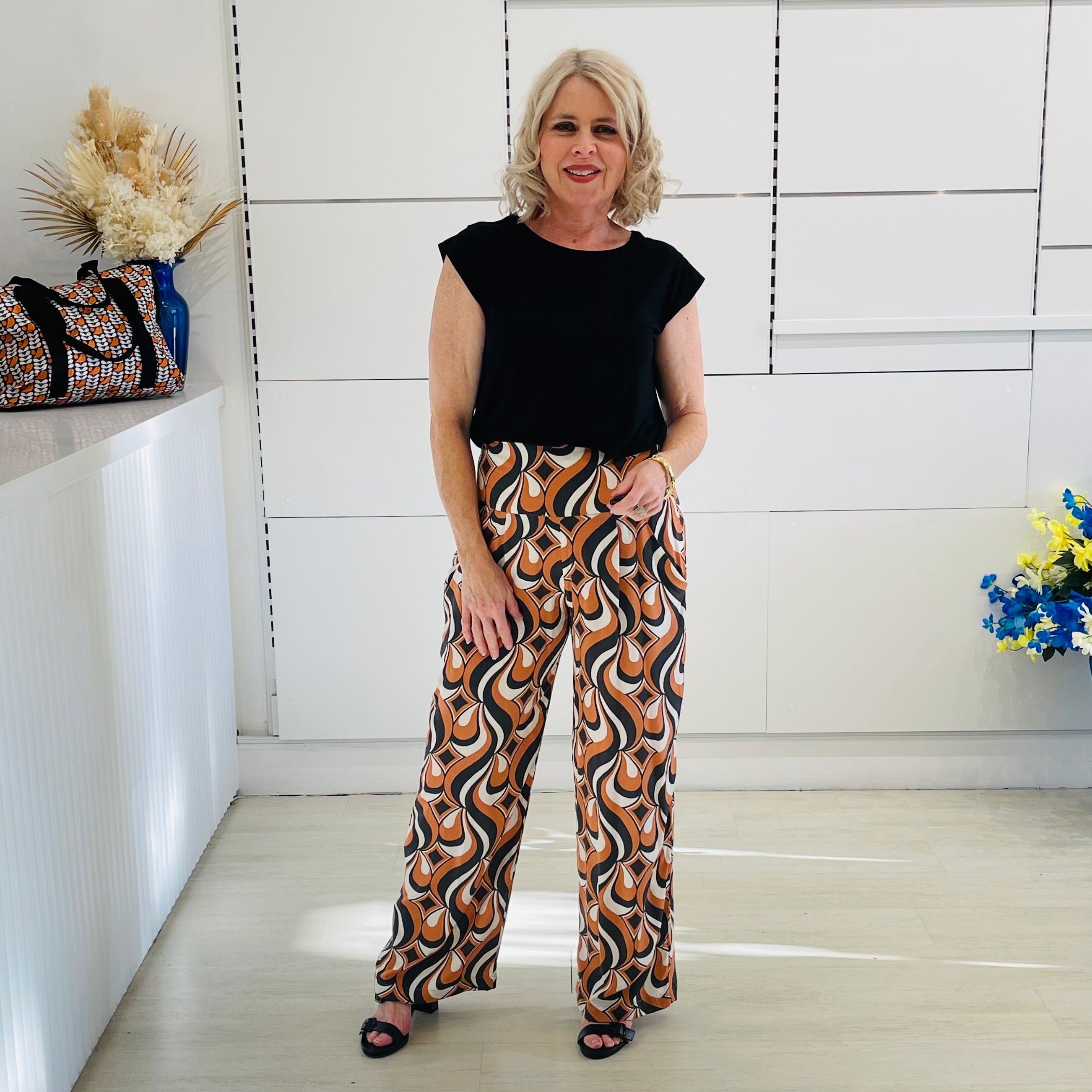Woman wearing a black top and patterned pants standing in a room with a white wall and decorative elements.
