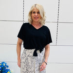 Woman wearing a black top and patterned pants standing against a white tiled wall.