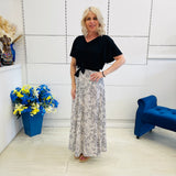Woman in a black top and patterned skirt standing in a room with a blue chair and flower arrangement.