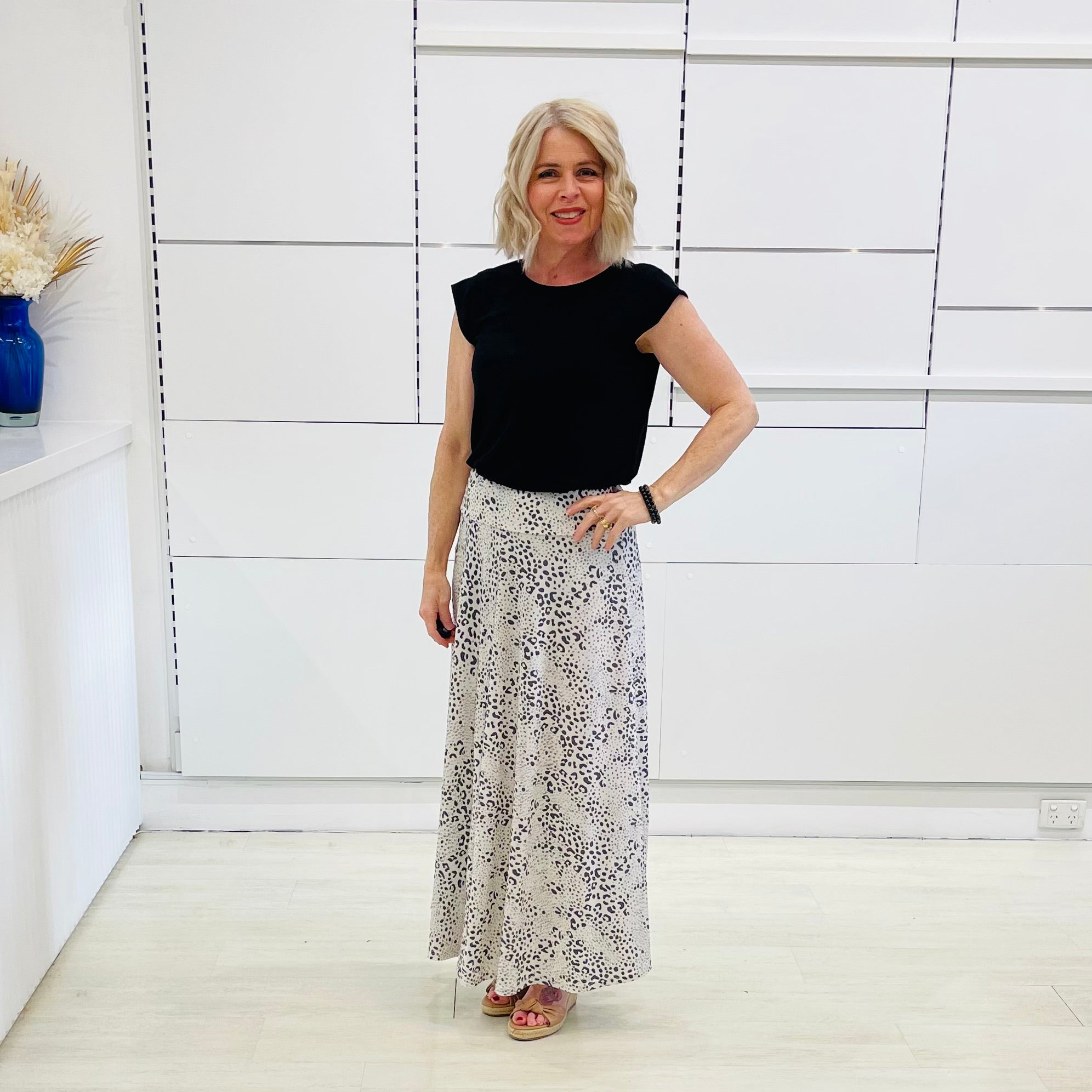 Woman wearing a black top and patterned skirt standing in a room with white walls and a shelf.