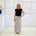 Woman wearing a black top and patterned skirt standing in a room with white walls and a shelf.
