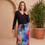 Woman wearing a black top and colorful skirt standing next to a potted plant against a beige wall.