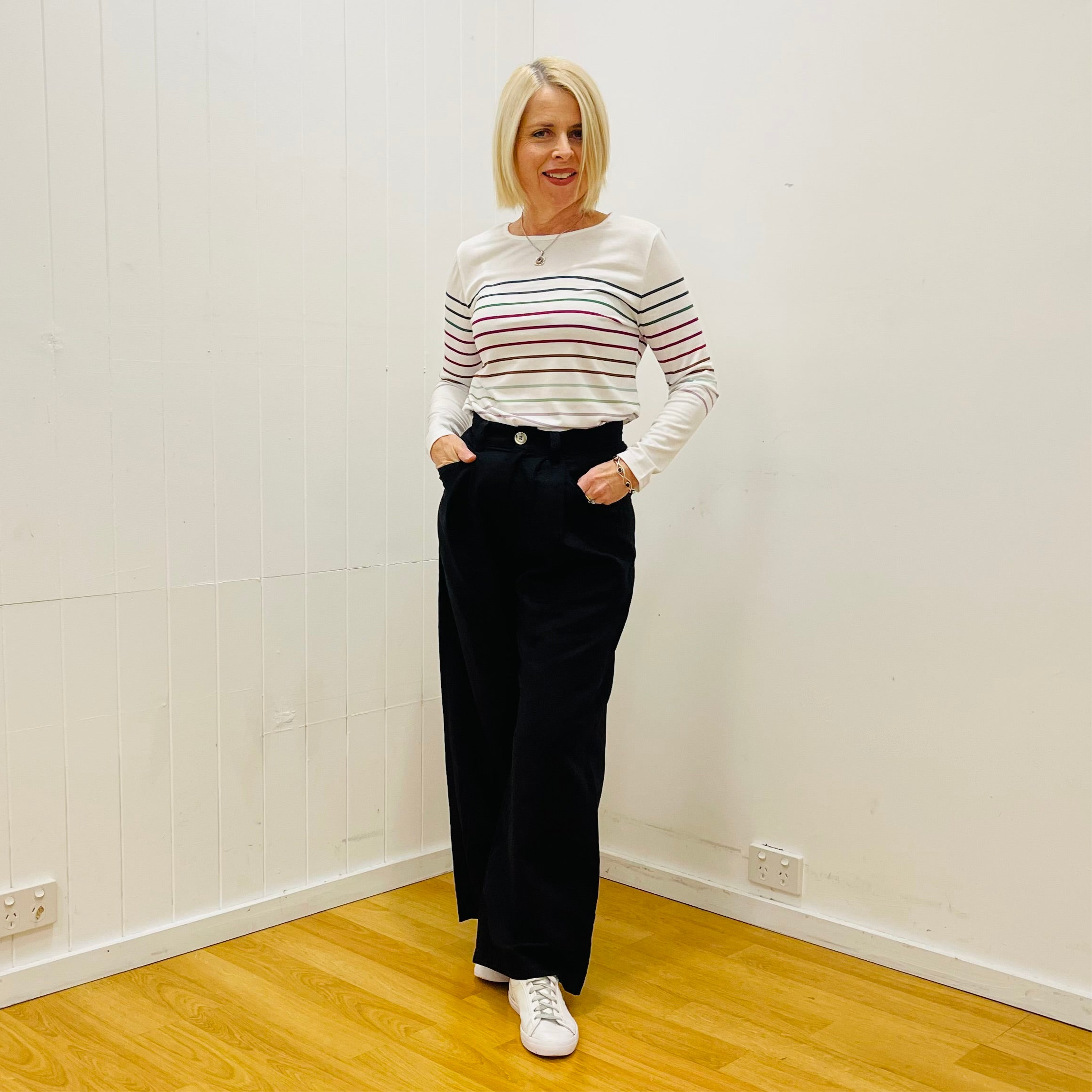 Woman wearing a striped sweater and black pants standing in a room with white walls and wooden floor.