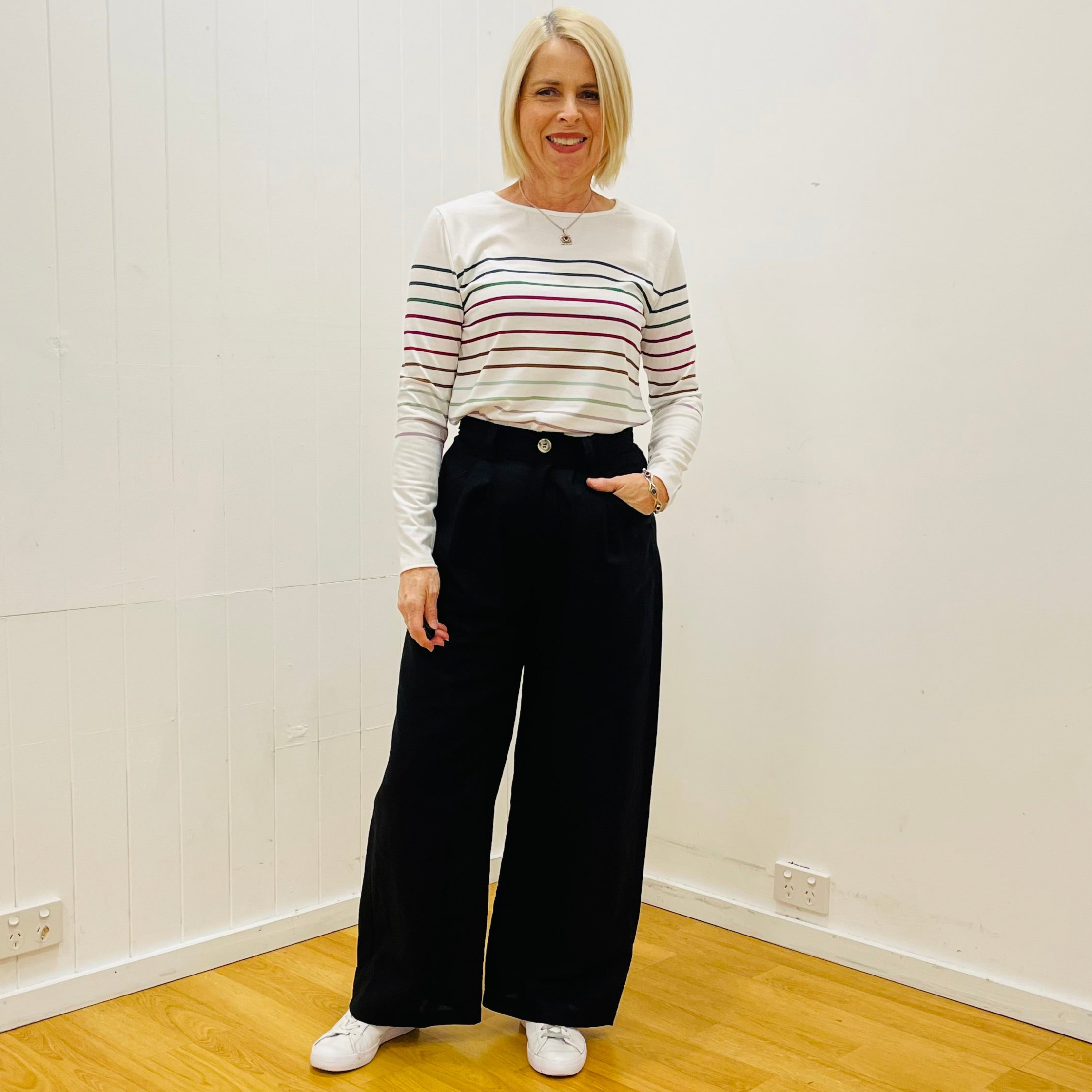 Woman wearing a striped shirt and black pants standing in a room with white walls and wooden floor.