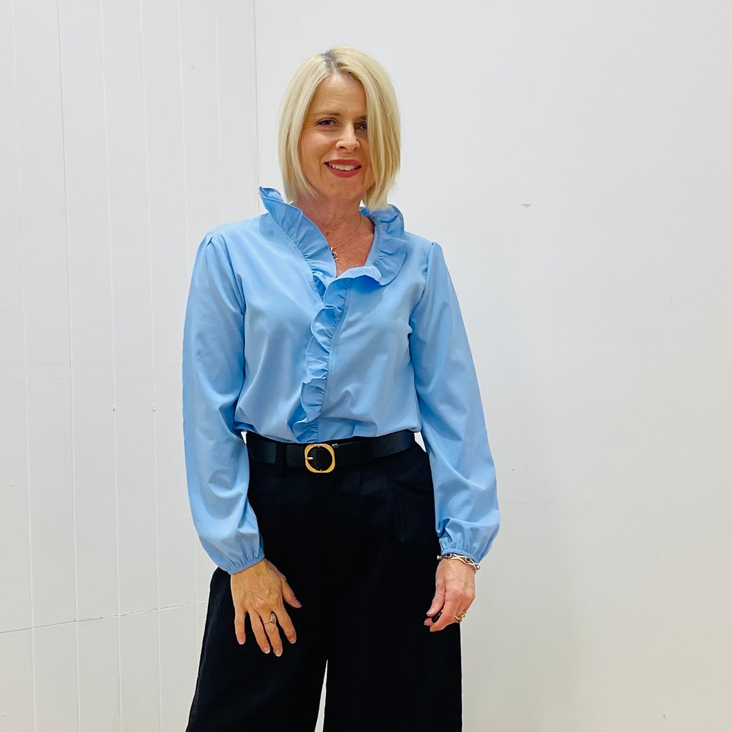 Woman wearing a light blue blouse with ruffled details and black pants against a white background