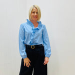 Woman wearing a light blue blouse with ruffled details and black pants against a white background
