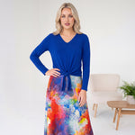 Woman wearing a blue top and colorful skirt in a room with a chair and plant.