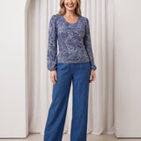 Woman wearing a blue patterned top and blue pants standing in front of a white curtain.