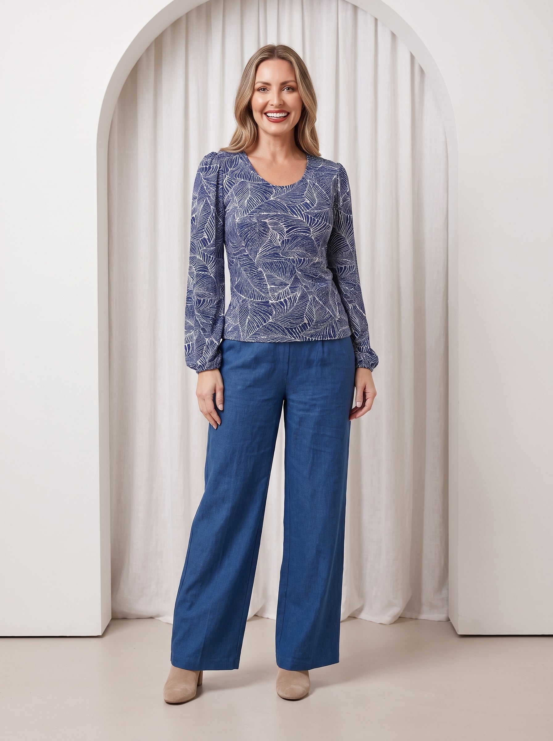 Woman wearing a blue patterned top and blue pants standing in front of a white curtain.