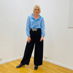 Woman wearing a light blue shirt and black pants standing in a room with white walls and wooden floor.