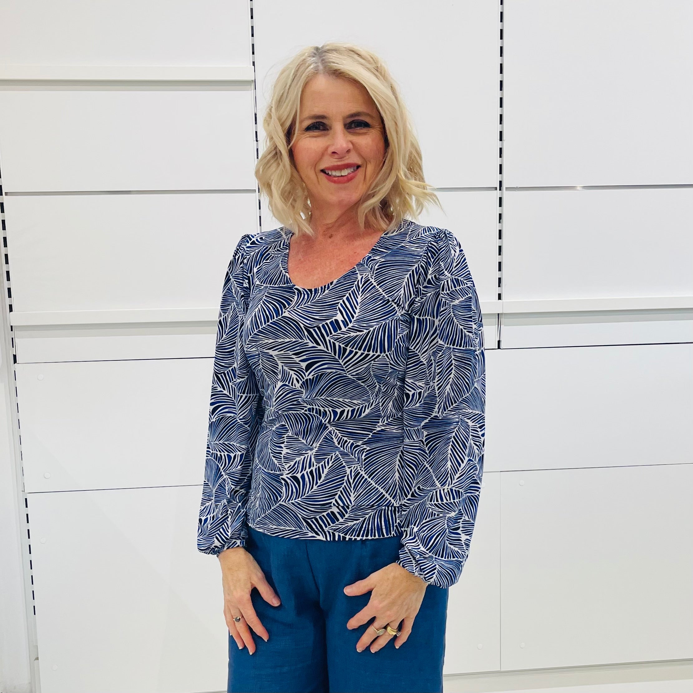 Woman wearing a scoop neckline, long sleeve,  flowy blue top with a delicate leaf print, paired with denim blue colored linen pants.