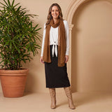 Woman wearing a white blouse, black skirt, brown scarf, and tan knee-high boots standing next to a potted plant against a beige wall.