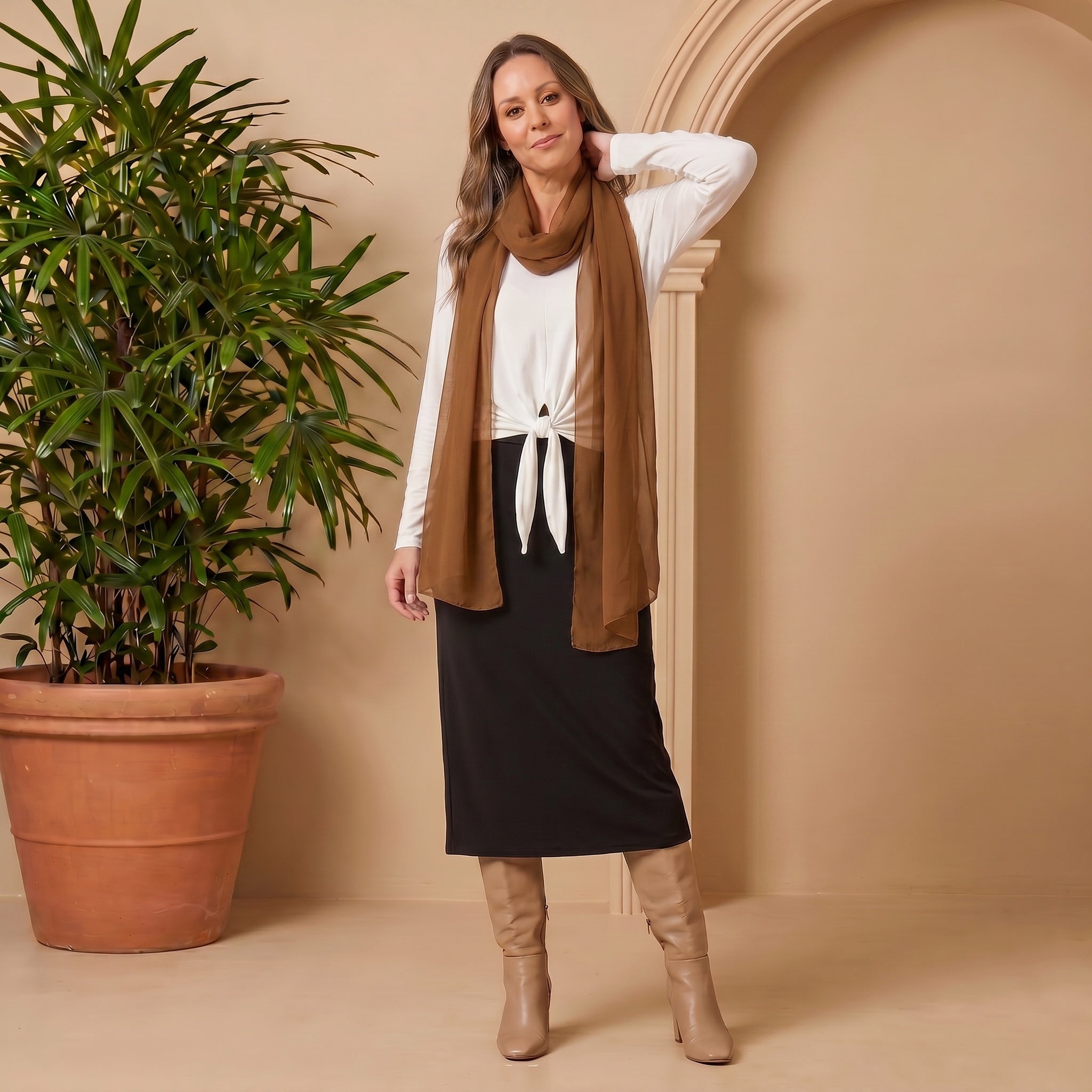 Woman wearing a white top, brown scarf, black skirt, and tan boots standing next to a potted plant against a beige wall.