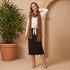 Woman wearing a white top, brown scarf, black skirt, and tan boots standing next to a potted plant against a beige wall.