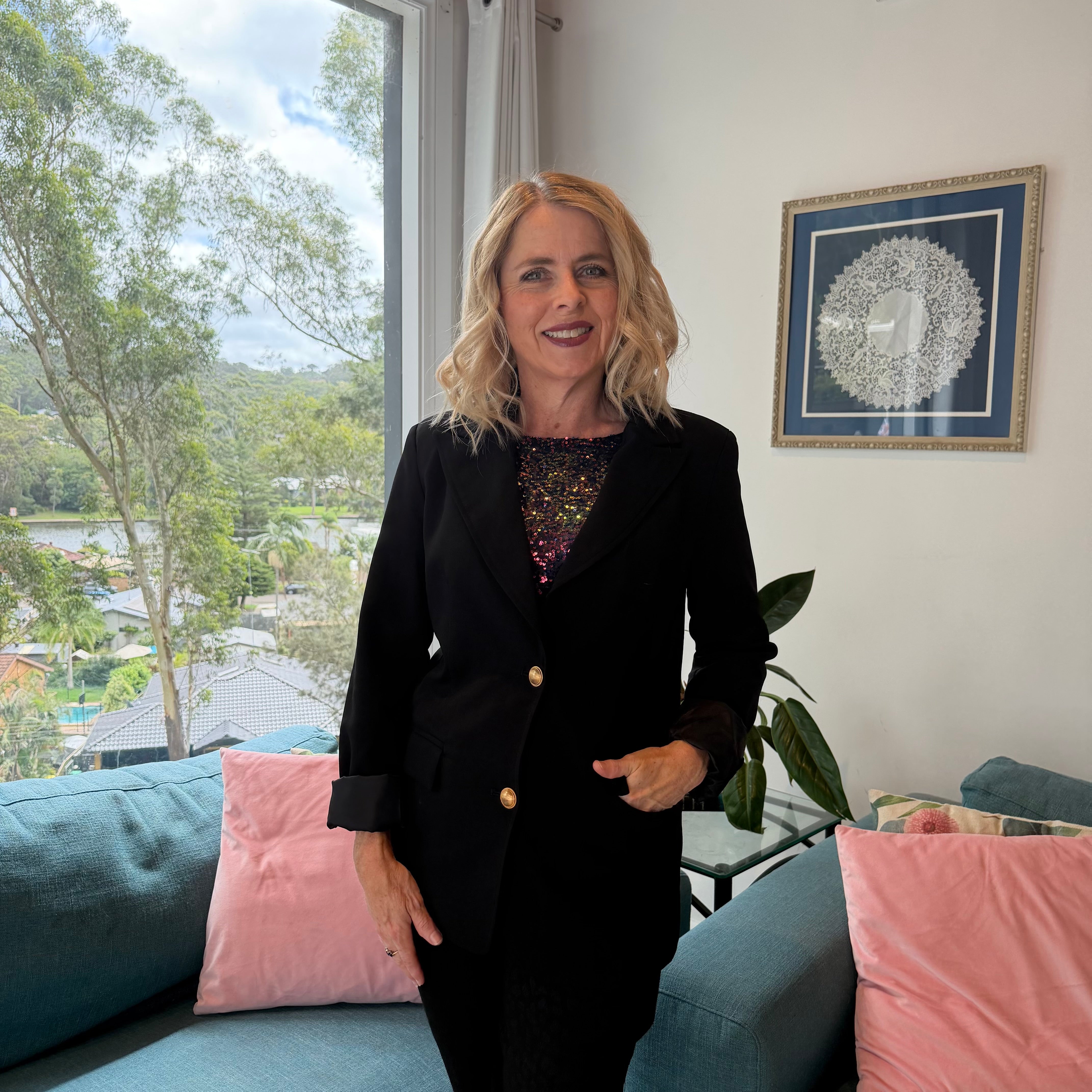Woman in a black suit standing in a room with a window view of trees and houses.