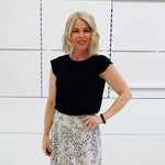 Woman wearing a black top and patterned skirt standing against a white wall.