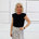 Woman wearing a black top and patterned skirt standing against a white wall.